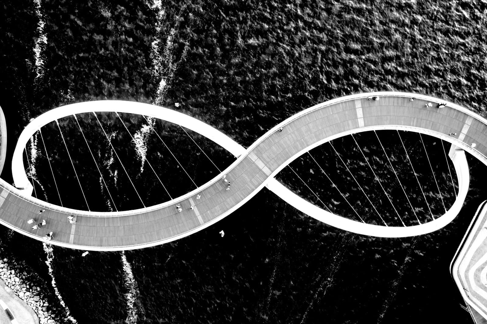 aerial view of a curved pedestrian bridge forming a figure‑eight shape over water, with people walking along it.