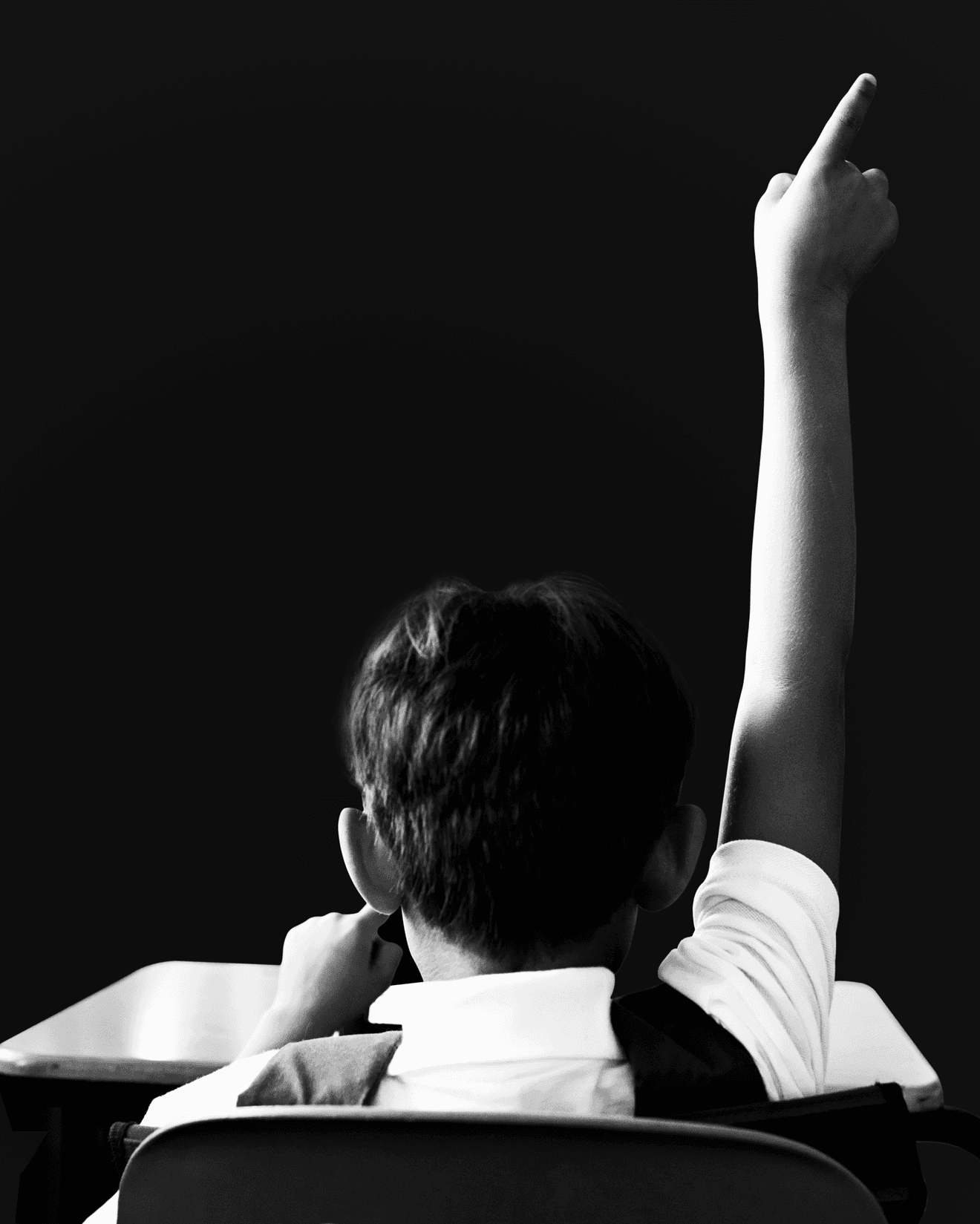 Child in white shirt and dark vest seated at desk with one arm raised against dark background.