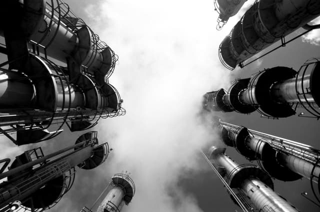 Industrial refinery towers viewed from below with clouds drifting overhead.